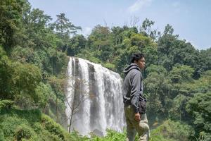 Man stand wear hoody in front of the great water fall with hand reel, Semarang central Java. The photo is suitable to use for adventure content media, nature poster and forest background.