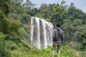 Man stand wear hoody in front of the great water fall with hand reel, Semarang central Java. The photo is suitable to use for adventure content media, nature poster and forest background.