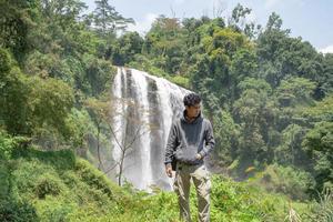 Man stand wear hoody in front of the great water fall with hand reel, Semarang central Java. The photo is suitable to use for adventure content media, nature poster and forest background.