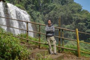 Man stand wear hoody in front of the great water fall with hand reel, Semarang central Java. The photo is suitable to use for adventure content media, nature poster and forest background.