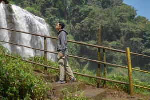 Man stand wear hoody in front of the great water fall with hand reel, Semarang central Java. The photo is suitable to use for adventure content media, nature poster and forest background.