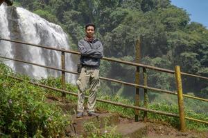 Man stand wear hoody in front of the great water fall with hand reel, Semarang central Java. The photo is suitable to use for adventure content media, nature poster and forest background.