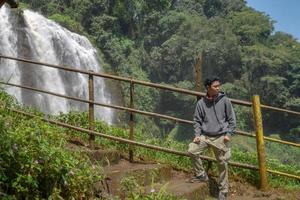 Man stand wear hoody in front of the great water fall with hand reel, Semarang central Java. The photo is suitable to use for adventure content media, nature poster and forest background.