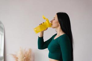 atlético joven mujer bebidas agua desde un amarillo botella después un rutina de ejercicio a hogar foto