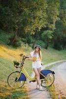 un joven hermosa mujer en pie siguiente a un bicicleta con un mimbre cesta lleno de flores en el bosque, participación un vaso de Fresco jugo con un Paja foto