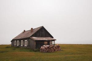 Lonely old wooden village house in heavy fog. Woodpile of firewood on green grass. Wind generator of electricity. Cloudy weather. photo