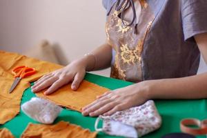 The seamstress smoothes the fabric blank for the mask. Fabric orange on a green table. Nearby are scissors and a finished mask. Focus on hands and tissue. Horizontal. photo