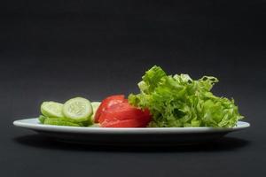 A photo of cucumbers, tomatoes and lettuce on a plate on isolated black background paper, after some edits.