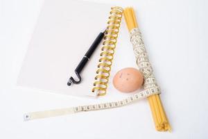 Bunch of raw spaghetti tied with measuring tape, chicken egg and notebook for writing with black pen on white background photo