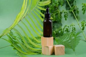 Serum glass bottle over wooden cubes with leaves on green background. Beauty product packaging display photo