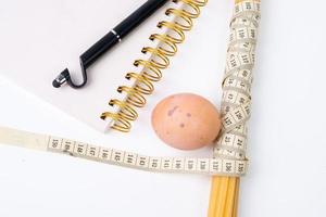 Bunch of raw spaghetti tied with measuring tape, chicken egg and notebook for writing with black pen on white background photo