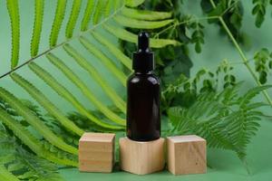 Serum glass bottle over wooden cubes with leaves on green background. Beauty product packaging display photo