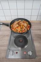 Electric stove in the kitchen with a frying pan cooking chicken legs and vegetables, with copy space background. Concept food and economic crisis photo