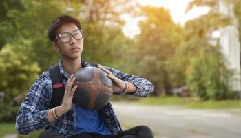 Portrait of asian male teenager in plaid shirt wear rainbow wristband and holding old and training basketball in hand with blurred park background, sunlight edited. photo