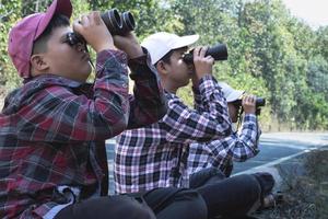 asiático Niños en tartán camisa usa gorra y participación un prismáticos, sentado en pavimento en local nacional parque a observar aves en árbol ramas y en cielo y a reloj insectos en árbol hojas. foto