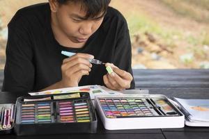 Asian boy is spending his free times during his summer vacation in the living room by drawing, painting and sketching with watercolor dry bar in tray in front of him, soft focus. photo