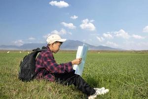 Asian boy wearing plaid shirt and a cap sitting on ridge of rice paddy field, holding a map and a binoculars, reading map before observing birds, pm 2.5 smoke and farmland borders, soft focus. photo