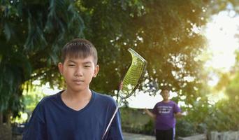 Young asian boy holding broken frame badminton racket in hand waiting to play badminton, soft and selective focus, outdoor badminton playing concept. photo