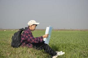 Asian boy wearing plaid shirt and a cap sitting on ridge of rice paddy field, holding a map and a binoculars, reading map before observing birds, pm 2.5 smoke and farmland borders, soft focus. photo