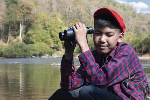 Asian boy in red cap and plaid shirt holds binoculars and sitting on rock in the middle of river in local national park to observe fish, nature and to watch birds on tree branches and flying on sky. photo
