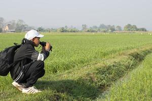 Asian boy wearing plaid shirt and a cap standing on ridge of rice paddy field, holding a map and a binoculars, looking through the lens to observe birds, pm 2.5 smoke and farmland borders, soft focus. photo