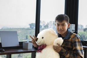 Asian cute boy in plaid shirt holding white bear doll, sitting near glass window in living room, ready to relax in his house, soft focus, concept for relaxing and recreational activity. photo