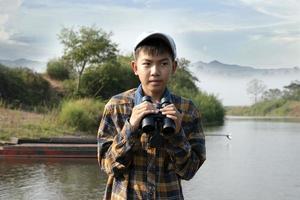 Asian boy in plaid shirt and cap, sitting and holding binoculars on bow of a boat parked beside a river to observe birds flying in the sky and fish swimming in the river during his summer holidays. photo