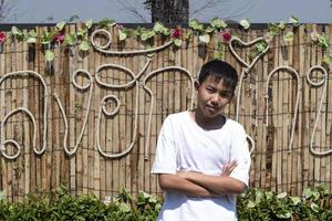 linda asiático chico en blanco camiseta, en pie con brazos doblada en frente de un bambú pared ese tiene tailandés caracteres en él, cuales medio 'en amar' en inglés. foto