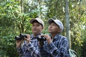 asiático Niños utilizando prismáticos a hacer el ornitología en tropical bosque durante verano acampar, idea para aprendizaje criaturas, fauna silvestre animales y insectos fuera de el aula. foto