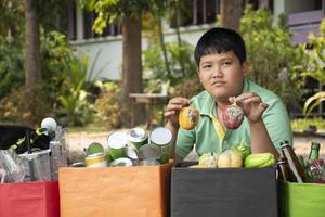 Asian boy is separating garbages and putting them into the boxes in front of him near building, soft and selective focus, environment care, community service and summer vacation activities concept. photo