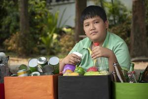 Asian boy is separating garbages and putting them into the boxes in front of him near building, soft and selective focus, environment care, community service and summer vacation activities concept. photo