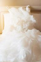 Wedding dress and suit lying on the white bed with beautiful sun light in hotel room. Valentines day and love for celebration concept. photo