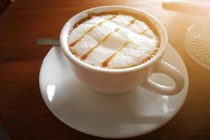 caliente café y espuma Leche en cerámico blanco taza con luz de sol en de madera mesa. Mañana hora foto