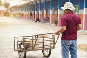 espalda ver de asiático hombre portero es que lleva rueda carro a mantener basura en escuela. concepto, Servicio ocupación. limpiar guardián para bueno ambiente. coleccionista basura a reciclar. Servicio hombre. foto