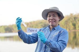 Asian woman ecologist hold tube of sample water and tag with word Type 4 for inspect and research water quality at the lake. Concept, explore, analysis water quality from natural source. Ecology field photo