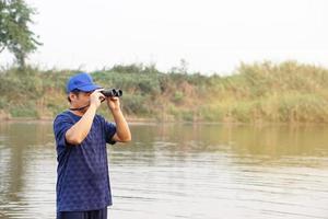 Asian man wears blue cap, hold binoculus to explore something at lake, nature source.Concept, nature exploration. Ecology study.  Pastime activity, lifestyle. Man explore environment photo