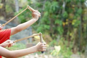 de cerca niños manos sostener honda a disparar planta semillas dentro bosque. concepto, creciente bosque por disparo tirachinas con semillas objetivo objetivo, caza o utilizar honda como armas o jugar juegos. foto