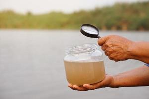 Closeup hands hold megnifying glass to inspect water in jar that contain sample water from the lake. Concept, explore, inspect quality of water from natural source. Ecology photo