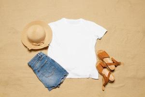 Sand beach texture background. Mockup white summer t-shirt copy space. Blank template woman shirt Top view. Summertime accessories hat, jeans, sandals. Flat-lay closeup tshirt seashore. Beachtime photo