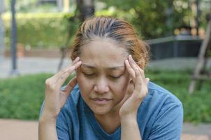 asiático mujer sentado a parque toque su templos, sensación un dolor de cabeza o mareado, sensación cansado. necesitar analgésicos foto