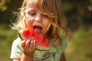 linda divertido pequeño niña comiendo un pedazo de sandía en el jardín en Hora de verano foto