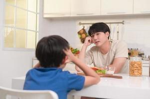 joven asiático padre enojado su hijo acerca de comer vegetales en cocina a hogar foto