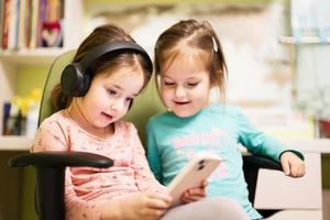 Two little girls sisters wear headphones watching cartoons or kid video on mobile phone. photo