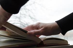 un persona leyendo libros cerca el ventana. manos vueltas terminado libro página. foto