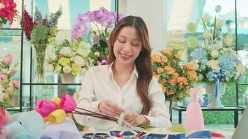 One Young beautiful Asian female florist entrepreneur arranging a bunch of blossoms, decorating with lovely ribbons, happy work in colorful flower shop store with blooms, and small business owner. video
