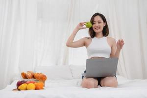 Portrait of a white Asian woman with tattoos sitting holding green apples and fruits in a healthy lifestyle. Relaxing in bed and playing on my laptop for the weekend. holiday concept. photo