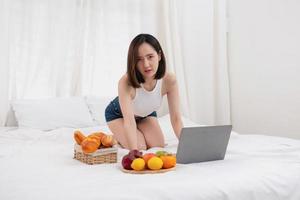 Portrait of white Asian woman with a tattoo sitting on the bed relaxing and playing laptop on weekend There was bread and fruit and red apples on a white bed to eat. holiday concept photo