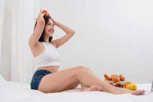 Portrait of white Asian woman with a tattoo sitting on the bed relaxing and playing laptop on weekend There was bread and fruit and red apples on a white bed to eat. holiday concept photo