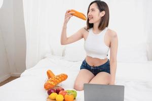 Portrait of a white Asian woman with a tattoo sitting and holding bread and fruit lying in bed eating happily. Relax in bed and play on your laptop on the weekends. holiday concept. photo