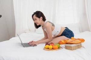 Portrait of white Asian woman with a tattoo sitting on the bed relaxing and playing laptop on weekend There was bread and fruit and red apples on a white bed to eat. holiday concept photo